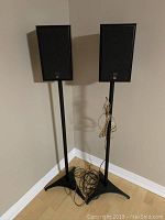 Pair of JBL 500 speakers mounted on black metal stands positioned in a corner on wooden floor. Cables are loosely placed at the base of the stands.
