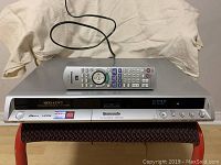 Front view of Panasonic DVD Recorder with remote placed on top showing power button, DVD slot, digital display, and various feature logos including HDMI and DIGA