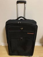 Front view of Renwick 29-inch black roller suitcase with extended handle, two zippered front pockets, Renwick metal logo, and an orange accent line on the upper pocket.