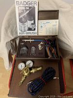 Open antique wooden case showing airbrush kit components arranged neatly inside foam insert including airbrush gun, jars, pipe cleaners, and manual.
