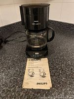 Philips black drip coffee maker with glass carafe, reusable filter inside, and instruction manual in front.