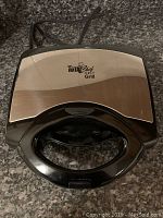 Closed view of the Total Chef 4-in-1 Grill/Panini Press on countertop showing black handle and stainless steel front plate with branding.