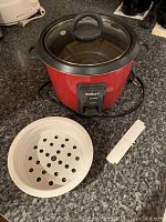 Red Saltor rice cooker on countertop with glass lid, white steaming basket and paddle in front.