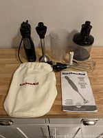 Full set of KitchenAid immersion hand blender components: motor unit, blending wand, whisk, food chopper, blending container, tote bag, and instruction manual.