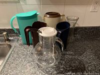 All six items arranged on kitchen counter: milk bag jugs, plastic pitchers, glass pitcher with ice core, and glass vase.