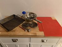 All items displayed on wooden countertop: black perforated BBQ tray, stainless steel food mill with blue crank handle, two red silicone mats, two thermometers (analog and digital), and extra grates for the food mill.