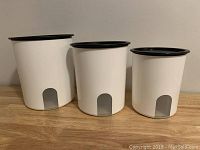Set of three white Tupperware containers with black lids, varying sizes lined up on a wooden surface, showing overall set condition and design.