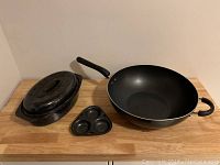 Large black wok, roasting pan with lid, and a small egg cooking pan displayed on wooden surface.