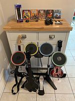 Overall display showing five PS3 game cases on countertop and three guitar controllers, drum kit, kick pedal, drum sticks, cables, USB adapters, and boxed PS3 controller on floor.