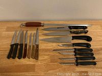 Photo of assortment of 15 kitchen knives and honing steel laid on wooden surface, showing two rows of knives with various blade types and handle materials