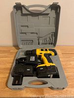 Open gray plastic case containing yellow and black 18V Power Fist cordless drill, spare battery, charger and drill/screw bits.