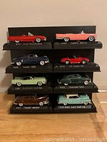 Front view of the eight vintage domestic car models mounted on a tiered black display stand with black display board, showing the vehicle names and Solido branding on the platforms.