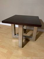 End table with dark wood square top and stainless steel geometric base on light wood floor, viewed from a side angle.