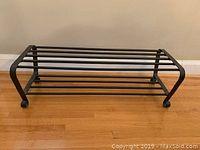 Photo of a black metal two-tier shoe rack on a hardwood floor against a beige wall.