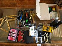 Photo showing clear plastic box filled with assorted hand tools including screwdrivers with different handles, pliers, wire cutters, smaller tools, ratchet set, flashlight, and a box with small hardware items.