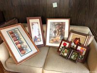 Eight framed artwork pieces displayed on a couch including several crewel embroidery pieces and prints.