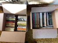 Top view of two cardboard boxes filled with various books, mostly paperbacks with some hardcover and larger format books mixed in