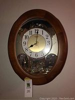 Front view of brown wood-tone oval clock on wall showing white dial, black Arabic numerals and decorative pendulum.