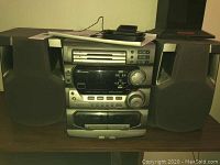 Front view of JVC MX-302T compact stereo system with two speakers, silver and black finish, showing CD player slots, buttons, dials and small display panel, placed on wood surface