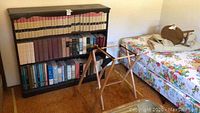 Wooden bookshelf filled with various hardcover books, foldable wooden luggage stand beside it, floral mattress background