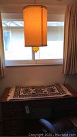 Vintage hanging light fixture with cylindrical fabric shade and amber hobnail glass orb seen lit near window.