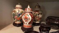 Three Japanese porcelain vases with intricate paint, each displayed on a separate wooden stand, showing front view and detailing.