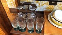 Wooden wine glass rack with four wine glasses, four glass coffee mugs with green handles, and some small glasses, plates, and trays on carpeted and wooden shelf surfaces.