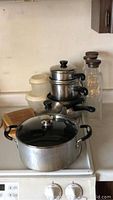Shows 5-quart metal stockpot with glass lid in front, three nested metal pots with black handles and lids behind, three glass containers stacked behind, and two wooden cutting boards stacked to the left.