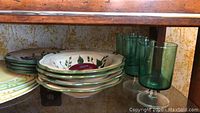 Dinner plates, small plates, bowls, and green glass goblets arranged on shelf under wooden table.