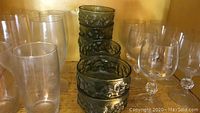 Various drink glasses on a wooden shelf: stack of 8 green embossed glasses, 6 clear wine glasses, 3 tall clear glasses