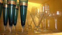 Front view showing six tall blue glasses with copper-colored bands and several assorted clear glasses on a wooden shelf.