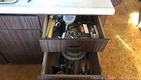Bottom drawer with assorted knives with wooden and plastic handles and other items such as cutting boards and plates.