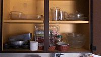 Open wooden cabinet with glass Pyrex bowls, casserole dishes, measuring cups, metal baking pans, white ramekins, and other kitchen items arranged inside.