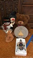 Photo showing two teddy bears, one snowman figure, and a decorative plate with a festive design, set on a carpeted floor in front of wooden furniture.
