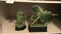 Two green plastic statues on black stands displayed on a shelf, showing front and side views.