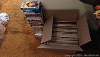 Photo showing a large open cardboard box filled with vinyl records and a tall separate stack of VHS tapes on a carpeted floor.