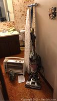 Photo showing Hoover Air vacuum upright, ironing board with floral cover, white portable heater, and a small iron on the floor in a room with carpeted floor and floral wallpaper.
