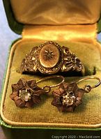 Gold colored vintage ring with oval face featuring clear stone and filigree detailing, displayed in green velvet ring box alongside matching flower earrings.