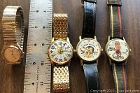 Four vintage wristwatches placed side by side next to ruler for scale, showing distinct dial designs and different band materials.