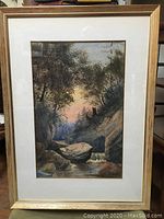 Framed watercolor showing full artwork including a woodland stream landscape with rocky terrain and sunset sky.