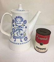 Full view of white ceramic tea pot with blue and green floral and figure design next to a vintage Campbell's soup can for scale.