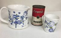 Two ceramic pitchers with blue, green, and purple floral and female figures next to a Campbell's soup can for scale.