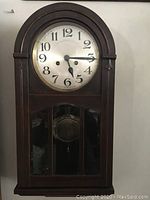 Front view of vintage wooden wall clock with arched top, beveled glass panel on front, clock face with black numbers and hands, and visible pendulum inside.