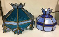 Two blue stained glass lampshades side by side on a surface, showing scale and distinct shapes.