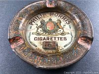 Full view of the round metal ashtray with colorful ornate rim and central advertisement for Philip Morris Cigarettes featuring a lion and unicorn coat of arms and cigarette box.