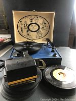 Blue portable record player with inside lid illustration and tone arm, vinyl record on spindle, Polaroid camera in front, one 45 RPM record on side