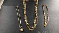 Three gold-colored vintage necklaces on black background showing different chain styles and pearl-like beads in one and a locket pendant on another.