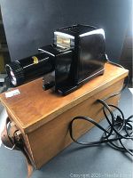 Chocolate brown Bakelite Kodak Kodaslide Merit projector placed on top of wooden storage case showing front lens and attached power cord with on/off switch.