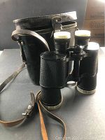Black leather binocular case with leather strap and vintage binoculars placed in front, showing textured grips and lens caps.