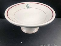 Front view of white ironstone pedestal cake stand with maroon band and green logo.